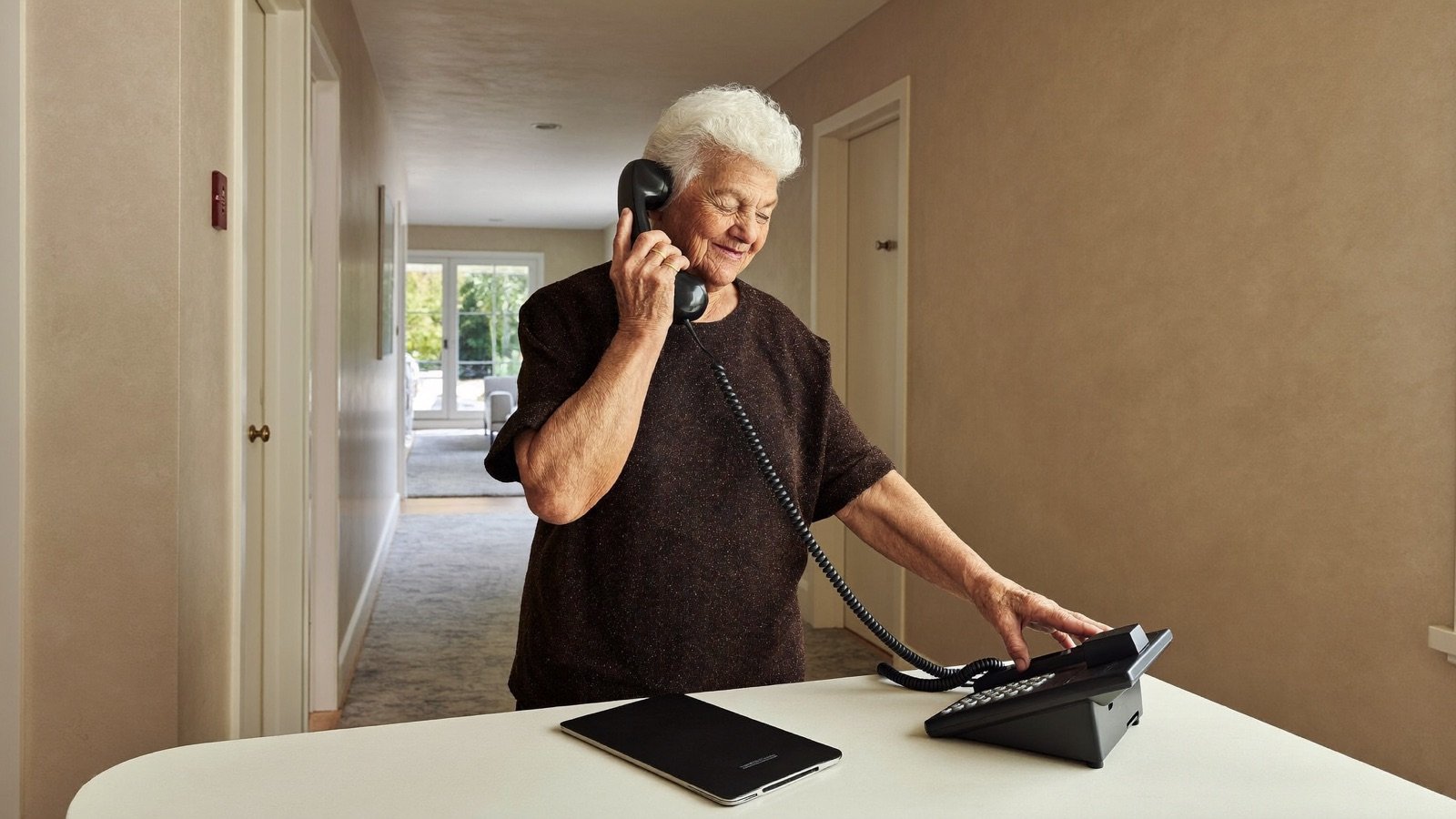 An older woman holding a black telephone receiver beside its base in a hallway.