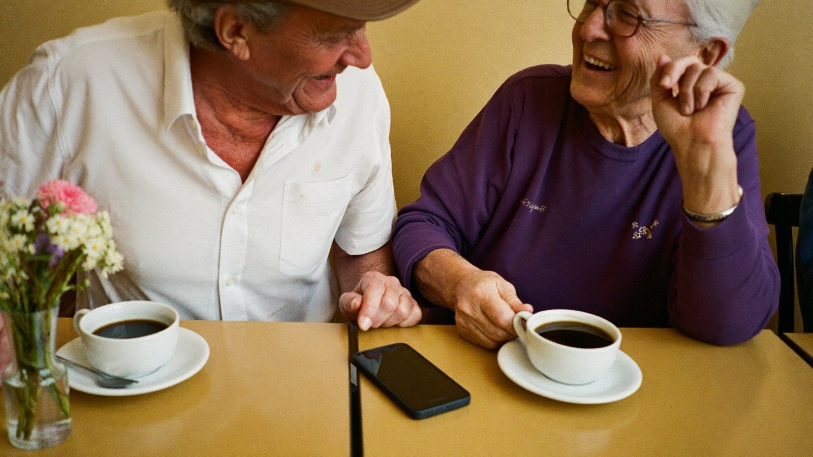 Two friends in their seventies compare their phones cheerfully over coffee.