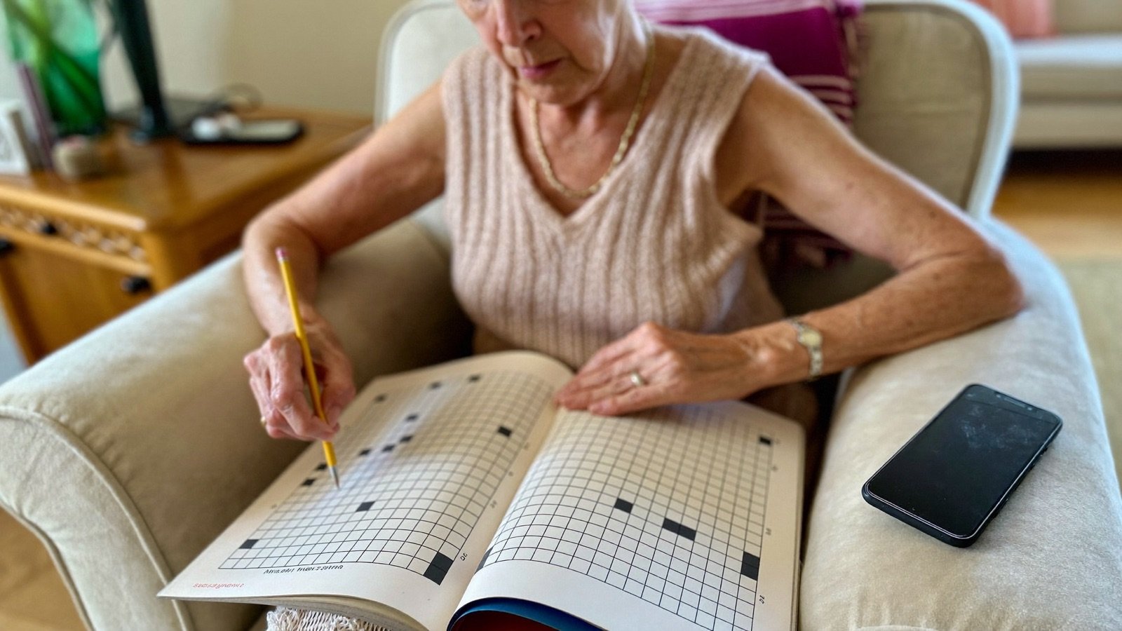 A woman in her eighties enjoys a crossword in a sunny armchair, phone resting nearby.