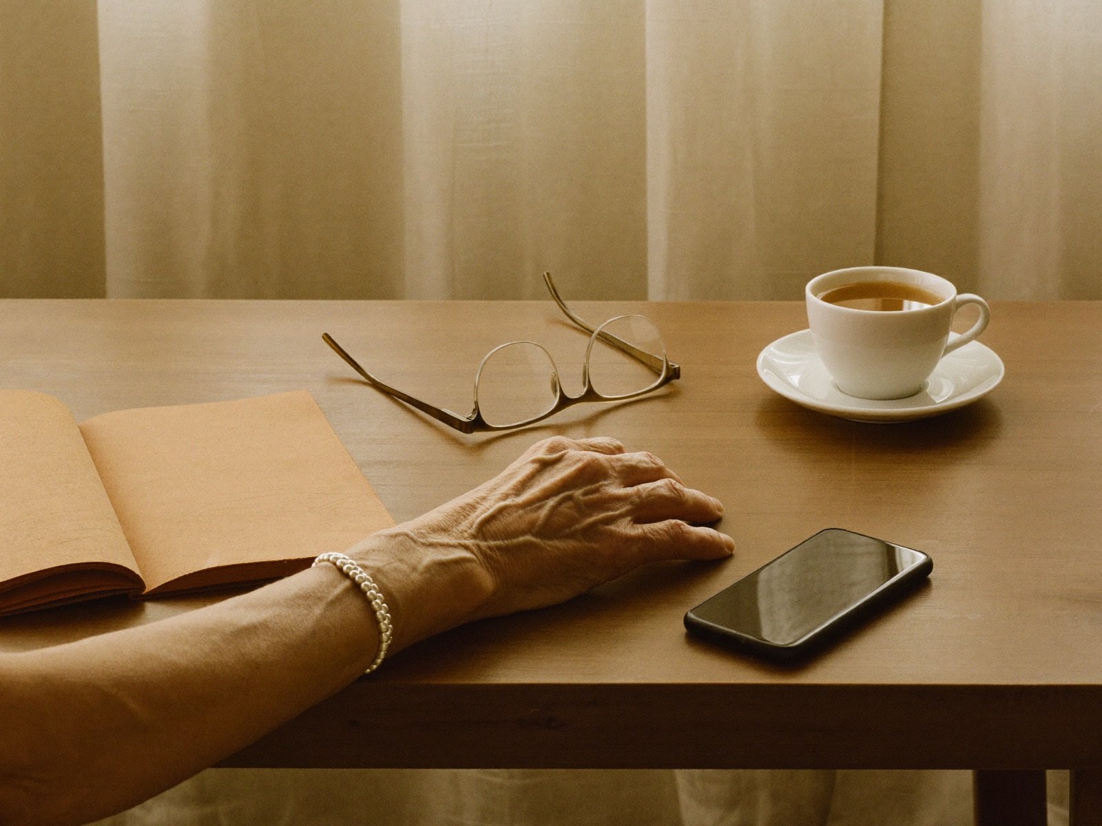 A calm still life: an older hand resting on a table beside an open notebook, reading glasses, a cup of tea, and a face-down phone in warm light.